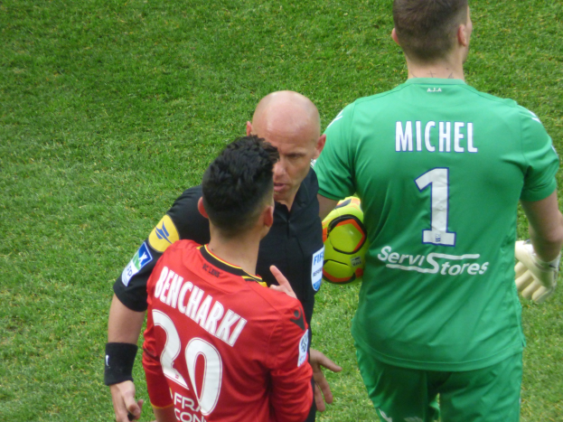 Ein Schiedsrichter spricht mit zwei Fußballspielern auf einem grasbewachsenen Feld, wobei einer der Spieler einen Ball hält.