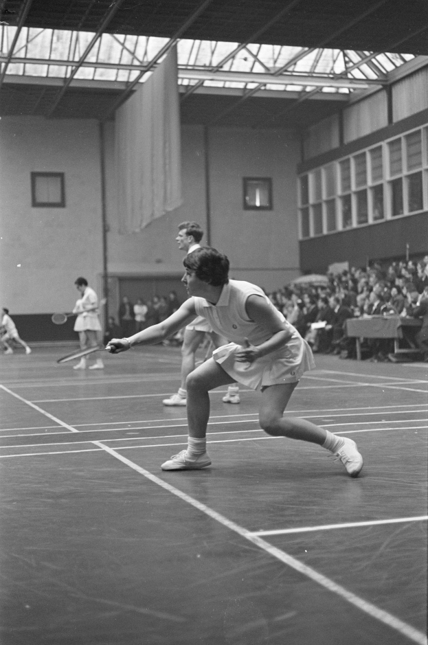Schwarzes und weißes Foto von zwei Frauen, die Badminton auf einem Platz spielen, mit Rackets in den Händen, umgeben von Zuschauern auf der rechten Seite und einer Wand mit Fenstern im Hintergrund.
