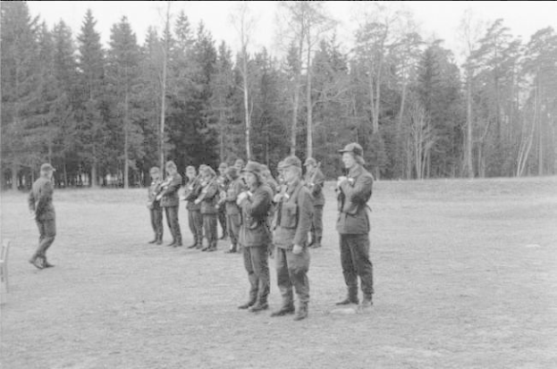 Schwarzes und weißes Bild einer Gruppe von Männern in militärischer Kleidung, die in einem Feld stehen und Gewehre halten, mit Bäumen und einem klaren Himmel im Hintergrund.