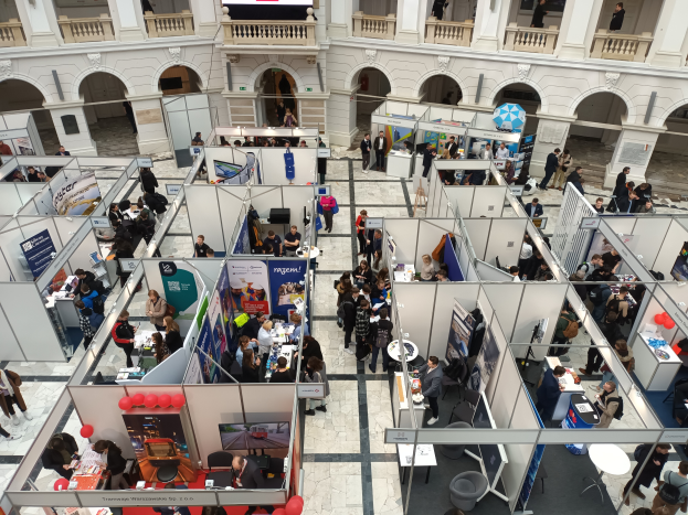 Eine große Gruppe von Menschen versammelte sich in einem Raum voller Stände, Banner, Tische, Stühle und Ballons, mit einem Gebäude mit Bögen, Säulen, Geländern und einem Bildschirm im Hintergrund.