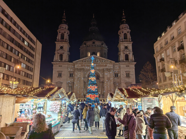 Ein belebter Weihnachtsmarkt vor einer Kirche bei Nacht, mit Menschen um geschmückte Stände, Gebäude, Laternen, Bäume und einen sternenklaren Himmel.