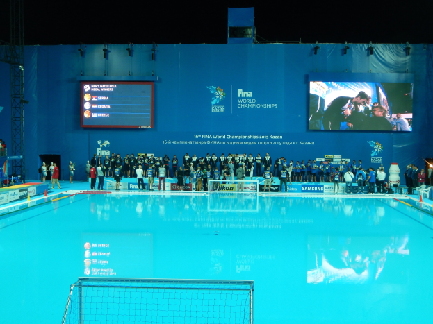 Gruppe von Menschen um ein Hallenbad mit Netz am Boden herumstehend, mit einer Wand im Hintergrund, die "FINA World Championships 2015 Kazan"-Text und Logos anzeigt, sowie Gerüste und Lampen