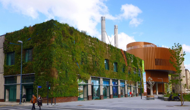 Großes nachhaltiges Gebäude mit einer grünen Wand, umgeben von städtischer Infrastruktur einschließlich Straßenlaternen, Ampeln, Fahrzeugen, Fußgängern, Bäumen und einem bewölkten Himmel.