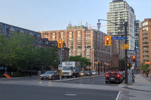 Eine stark befahrene Stadtstraße mit einem Lastwagen, einem Auto und einem SUV, gesäumt von Laternen, Ampeln, Anzeigetafeln, Bäumen und Gebäuden vor einem Himmel.