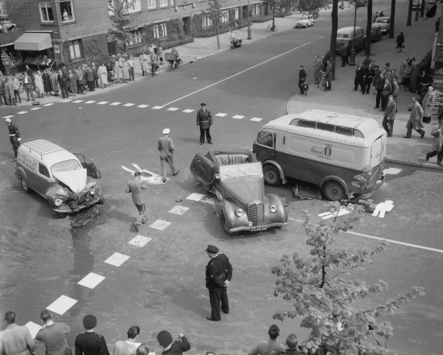 Schwarz-weißes Bild einer Autounfall-Szene an einer Straßenkreuzung mit mehreren Fahrzeugen, darunter ein Van, und einer Gruppe von Schaulustigen; Gebäude, Bäume, Pfosten und Texttafeln im Hintergrund.