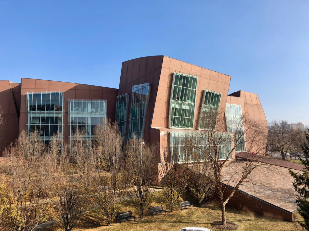 Großes modernes Universitätsgebäude mit vielen Fenstern, umgeben von Bäumen, Bänken, Gras und einem Laternenmast, mit anderen Gebäuden und einem klaren blauen Himmel im Hintergrund.