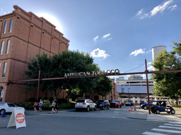 Eine belebte Stadtstraße mit Fahrzeugen, Fußgängern, einem Schild, Verkehrskegeln, Bäumen, Gebäuden, einem Bogen mit Text und Wolken, mit der American Tobacco Factory in St. Louis, Missouri in der Mitte.