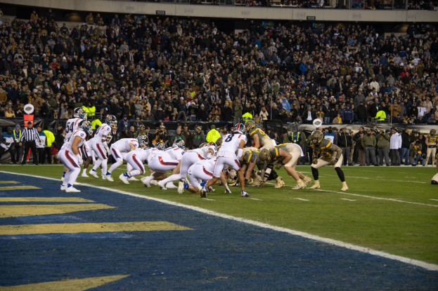 Fußballspieler in dunklen Uniformen und Helmen liegen auf dem Feld während eines Spiels, mit Zuschauern und Stadionelementen im Hintergrund.
