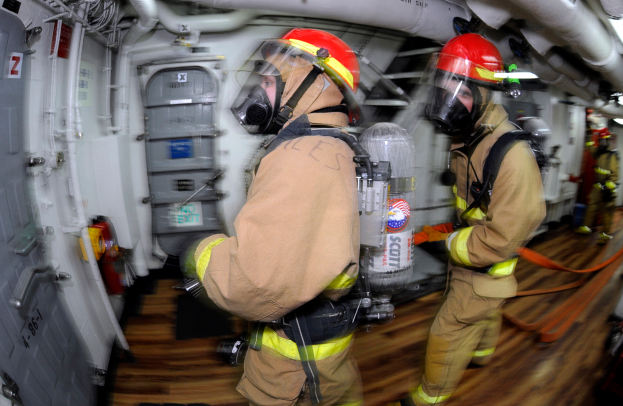 Eine Schwarz-Weiß-Zeichnung einer chaotischen Theater-Szene mit Menschen in Not, einige schauen ängstlich nach oben und andere verzweifelt nach unten, während das Theater in Flammen steht und Rauch aufsteigt.