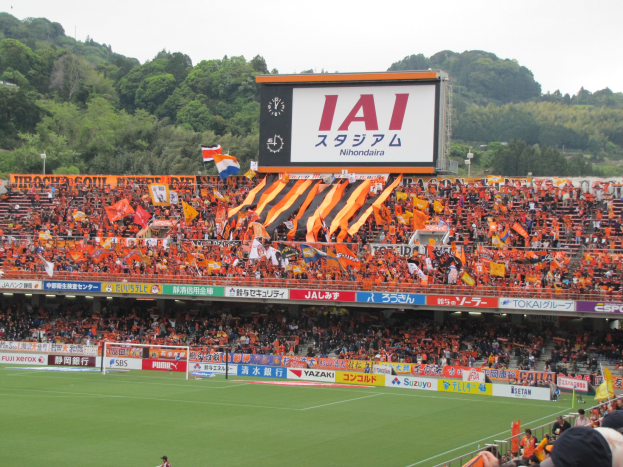 Ein Fußballspiel wird in einem Stadion mit einer großen Zuschauermenge, grünem Rasen, einem Torpfosten, Bannern, Fahnen, einem großen Bildschirm, Bäumen und einem klaren blauen Himmel gespielt.