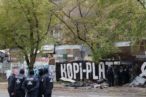Eine Gruppe von Polizisten in schwarzen Uniformen und Helmen steht vor einem mit Graffiti übersäten, baufälligen Gebäude mit verstreuten Trümmern und Bäumen im Hintergrund.