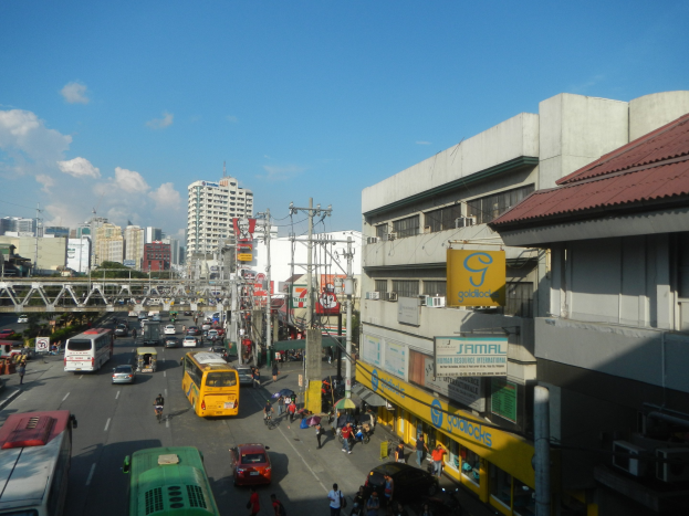 Eine belebte Stadtstraße mit zahlreichen Fahrzeugen, Fußgängern auf den Gehwegen, Strommasten mit Kabeln, Gebäuden mit Namensschildern, eine entfernte Brücke, Bäume und ein bewölkter Himmel.