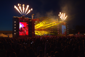 Eine große Menschenmenge versammelte sich auf einem Musikfestival mit einer Bühne in der Mitte, auf der Musiker Instrumente spielten, beleuchtet von hellen Lichtern und umgeben von Bannern, Zelten links und einem dunklen Himmel mit Feuerwerk im Hintergrund.
