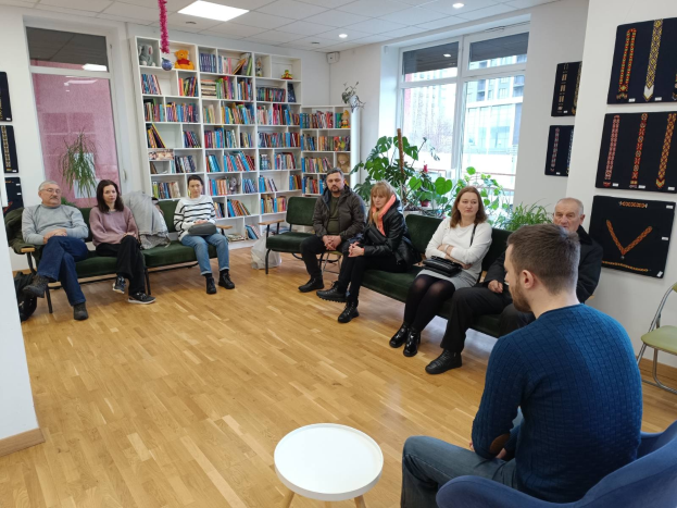 Gruppe von Menschen, die um einen Tisch in einem Raum mit Bücherregalen, Pflanzen und Deckenleuchten sitzen und während einer Workshop-Diskussion über die Zukunft von Gesundheit und Wohlbefinden in Berlin, Deutschland, diskutieren.
