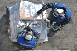 Zwei Männer in blauen Uniformen und Helmen arbeiten an einem großen Drohnenmodell, hältend verschiedene Gegenstände, mit einer Plastikabdeckung vor ihnen auf dem Boden.