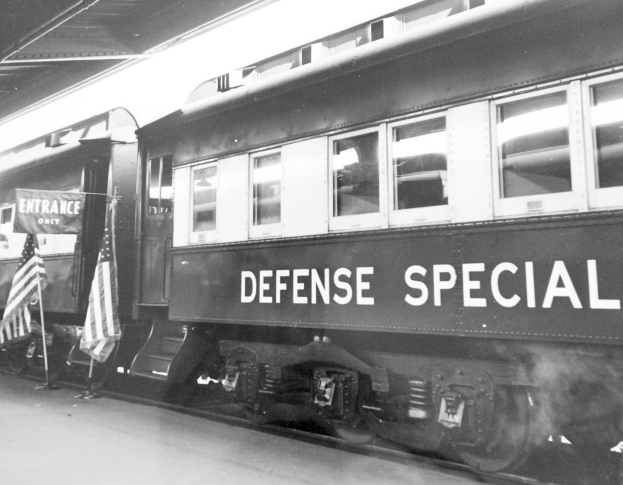 Schwarz-weißes Foto eines Zuges mit der Aufschrift "Defense Special" auf einem Gleis mit zwei Fahnen links und einer Decke oben.