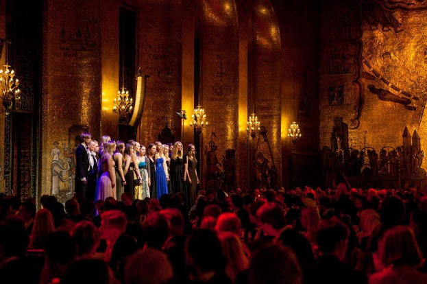 Eine Gruppe von Menschen auf einer Bühne im Royal Opera House in London, mit einem Publikum darunter, unter Bühnenbeleuchtung und Wandgemälden.