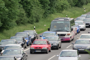 Stau auf einer Autobahn mit vielen Autos und einem Lieferwagen, Menschen in den Fahrzeugen sichtbar, mit Bäumen und Gras im Hintergrund.
