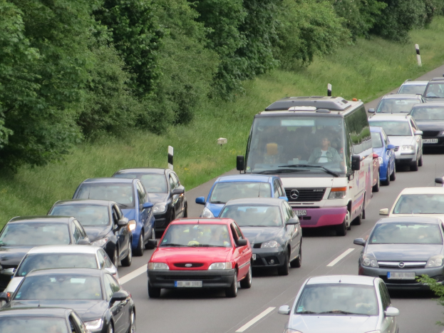 Stau auf einer Autobahn mit vielen Autos und einem Lieferwagen, Menschen in den Fahrzeugen sichtbar, mit Bäumen und Gras im Hintergrund.