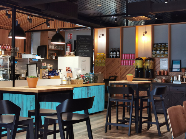 Innenraum eines Restaurants mit hölzernen Tischen und Stühlen, einem Blumentopf mit einer Pflanze darauf, einer Tafel, einem Kühlschrank, einem Feuerlöscher, Regalen mit Flaschen, Schränken mit Gegenständen, einem Rahmen mit einem Foto an der Wand und Deckenleuchten.