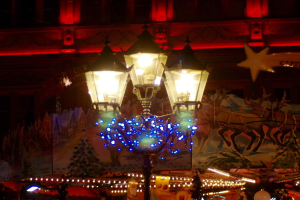 Eine mit Lichtern und festlichen Accessoires geschmückte Straßenlaterne steht nachts in einer Stadt, im Hintergrund ist ein Gebäude zu sehen.