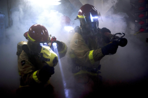 Zwei Feuerwehrleute in Schutzausrüstung sprühen Wasser auf eine rauchgefüllte Wand, mit einer Tafel mit Text auf der linken und verschiedenen Gegenständen auf der rechten Seite.