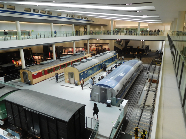 Ein großzügiger Museumsausstellungsraum zeigt verschiedene Züge in verschiedenen Größen und Farben, mit Menschen, die vom Boden, Geländern und Säulen unter Deckenbeleuchtung beobachten.