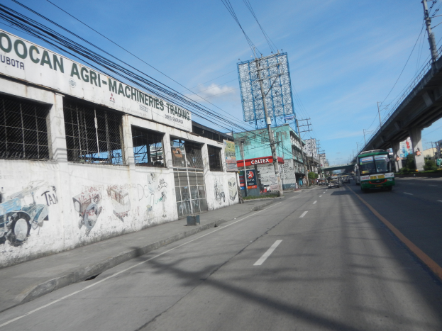Eine Straße mit fahrenden Fahrzeugen, eine Brücke auf der rechten Seite, Gebäude, Strommasten mit Käblein und ein bewölkter Himmel, mit einem graffiti-bedeckten Gebäude an der Straßenseite.