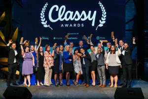 Eine Gruppe von Menschen auf einer Bühne, die strahlen und Auszeichnungen halten, mit Lautsprechern unten und einem Hintergrundbild mit der Aufschrift "Classy Awards 2019".