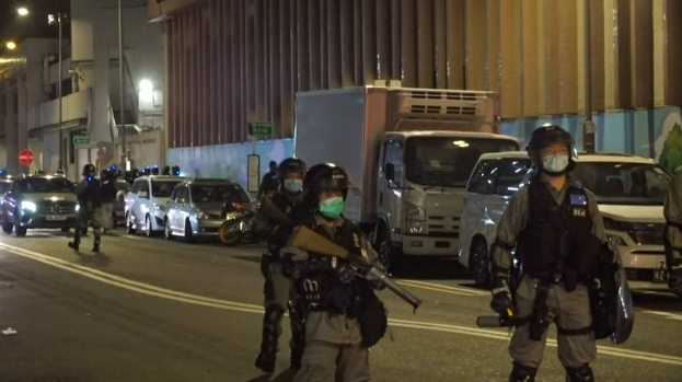 Eine Gruppe von Polizisten in Helmen, Handschuhen und Gesichtsmasken steht nachts auf einer Straße, einige halten Waffen, mit Fahrzeugen, Laternenpfählen, Schildern und Gebäuden im Hintergrund.