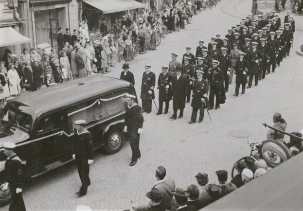 Schwarzer und weißer Trauerzug mit einem Leichenwagen, der den Körper eines Mannes trägt, umgeben von Menschen mit Waffen und Fahrzeugen, mit Gebäuden und Schildern im Hintergrund.