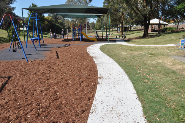 Ein Park mit einem Spielplatz mit Schaukeln und Rutschen, umgeben von grünem Gras, Bäumen, Bänken, Schuppen und einem klaren blauen Himmel.