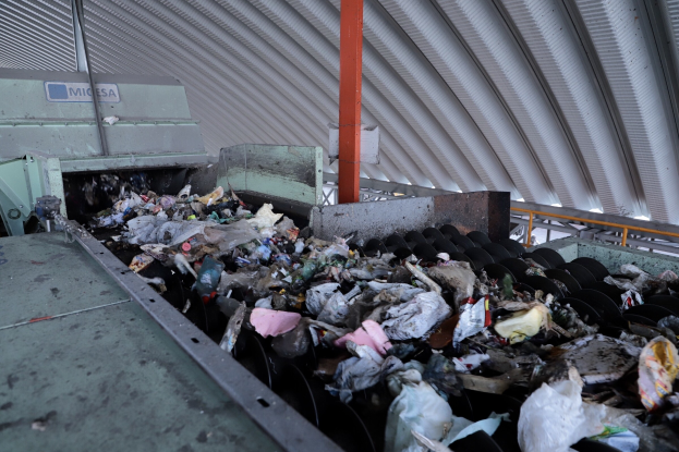 Großer Müllhaufen auf einem Förderband in einer Recyclinganlage mit einer Maschine und einem Metalldach im Hintergrund.