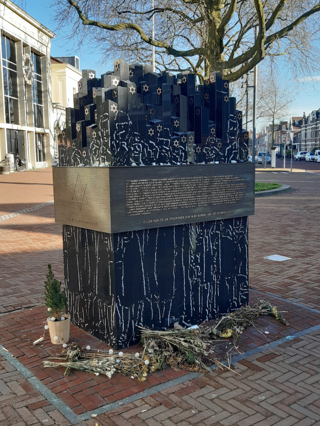 Denkmal für Holocaust-Opfer in einem Stadtplatz mit einem Baum davor, einem Topf mit einer Pflanze, verstreuten trockenen Blättern und Gebäuden, Fahrzeugen und Bäumen im Hintergrund.