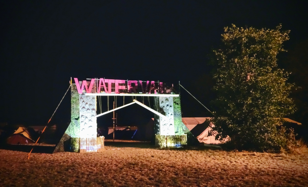 Bühne für ein nächtliches Musikfestival mit einem Tor mit Text, einem Baum auf der rechten Seite, Zelten im Hintergrund und einem dunklen Himmel.