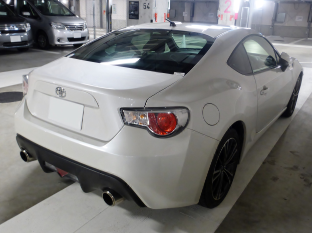 Ein weißer Sportwagen auf einem hell beleuchteten Parkplatz neben anderen Fahrzeugen geparkt, mit einer Wand im Hintergrund.