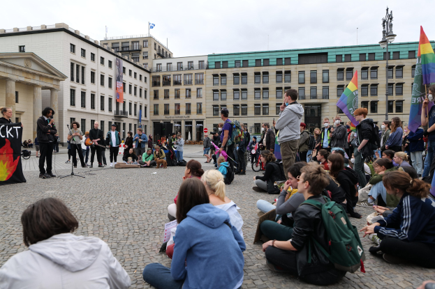 Eine Gruppe von Menschen, die auf dem Boden vor einer Menge sitzen, die Fahnen, Schilder und einen Mikrofonständer hölt, während einer Anti-Schwulen-Demo in Berlin, mit einer Statue, Gebäuden und einem bewölktem Himmel im Hintergrund.