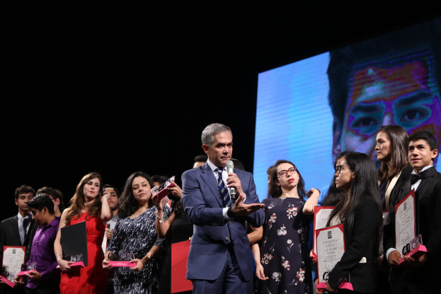 Eine Gruppe von Menschen auf einer Bühne, mit einem Mann in Anzug und Krawatte in der Mitte, der ein Mikrofon hält, umgeben von Personen, die Auszeichnungen halten, und einem Bildschirm im Hintergrund.