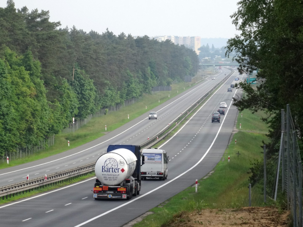 Ein großer Lkw fährt auf einer von Bäumen gesäumten Autobahn, die von einem Zaun und Gras auf der rechten Seite begrenzt ist, mit Gebäuden in der Ferne unter einem klaren blauen Himmel.