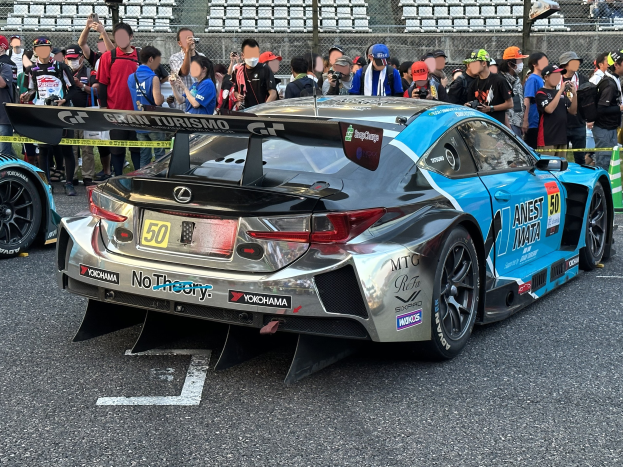 Ein schwarzer Lexus RC F GT3 Rennauto umgeben von Zuschauern mit Kameras und Handys, mit Maschendrahtzaun und verstreuten Stühlen im Hintergrund.