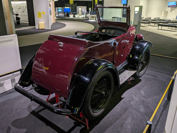 Ein klassisches rotes Auto in einem Museum ausgestellt mit Informationsplakaten, Barrieren und Sitzgelegenheiten.