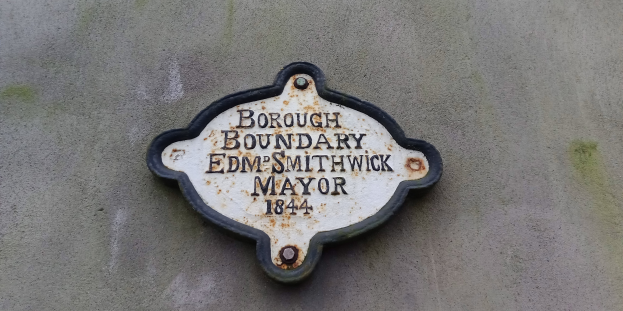 Schwarze Metalltafel an einem Gebäude mit weißer Schrift, die "Stadtteilgrenze Edm Smithwick Bürgermeister" in fetter Schrift lautet.