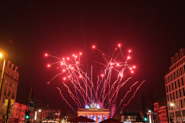 Eine Straßenstraße in Berlin an Silvester, voller Gebäude, Bäume, Laternenpfähle, Verkehrszeichen, Zelte und Menschen, mit einem Feuerwerk, das den Himmel erhellt.