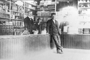 Ein Schwarz-Weiß-Foto von drei Männern, die an einem Tresen in einem Laden stehen, mit verschiedenen Gegenständen auf dem Tresen und einem Regal, das mit Büchern und anderen Gegenständen gefüllt ist, während eines Raubüberfalls.