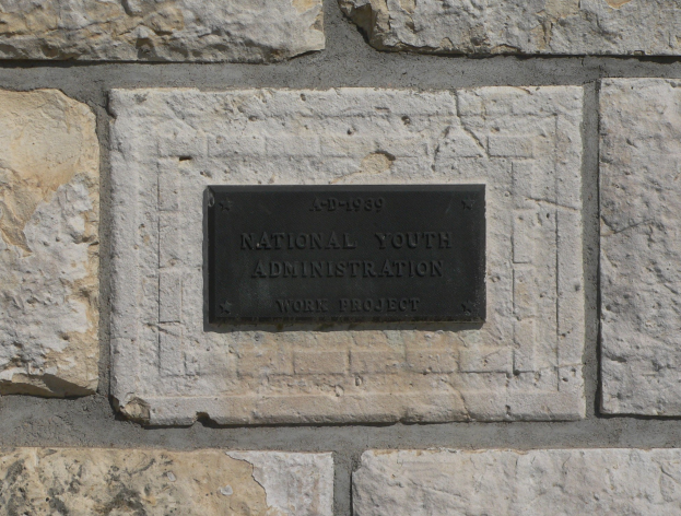 Steinwand mit einem rechteckigen Schild, auf dem "Nationaler Jugenddienst" in weißer, fetter Schrift auf schwarzem Hintergrund steht.