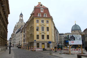 Eine Stadtstraße mit einem gelben Gebäude in der Mitte, umgeben von Straßenlaternen, Fahrzeugen, Fußgängern, Werbetafeln, Bäumen und einem klaren blauen Himmel.