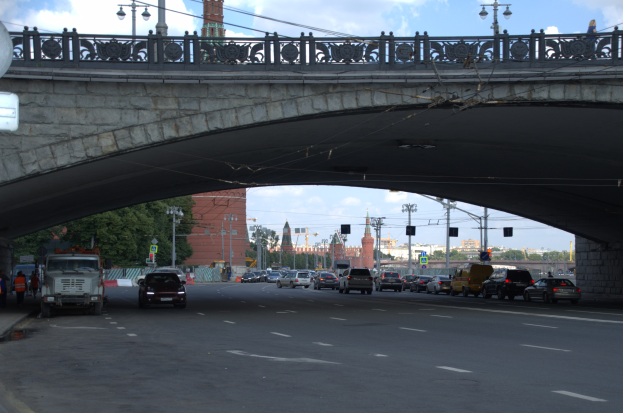 Eine Brücke überspannt eine belebte Straße mit darunterfahrenden Fahrzeugen, während Fußgänger auf dem Gehweg stehen; Gebäude, Bäume, Laternen, Verkehrszeichen und der Himmel sind im Hintergrund zu sehen.