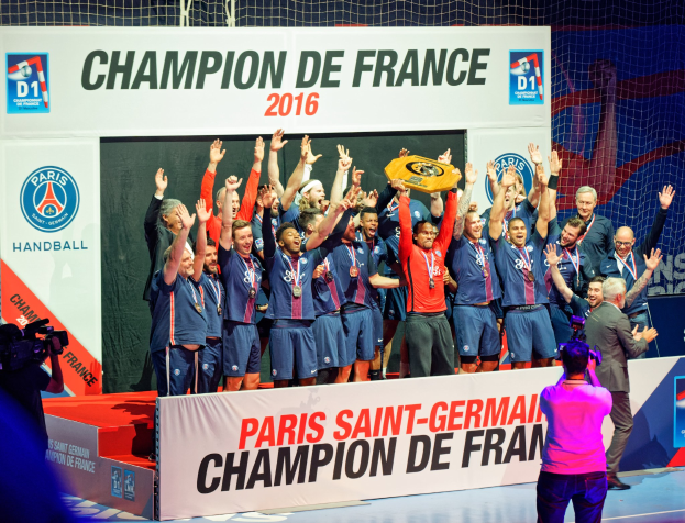 Eine Gruppe von Männern hält einen Pokal auf der Bühne, während eine Videokamera aufnimmt und ein "Champion de France 2016"-Schild im Hintergrund zu sehen ist.