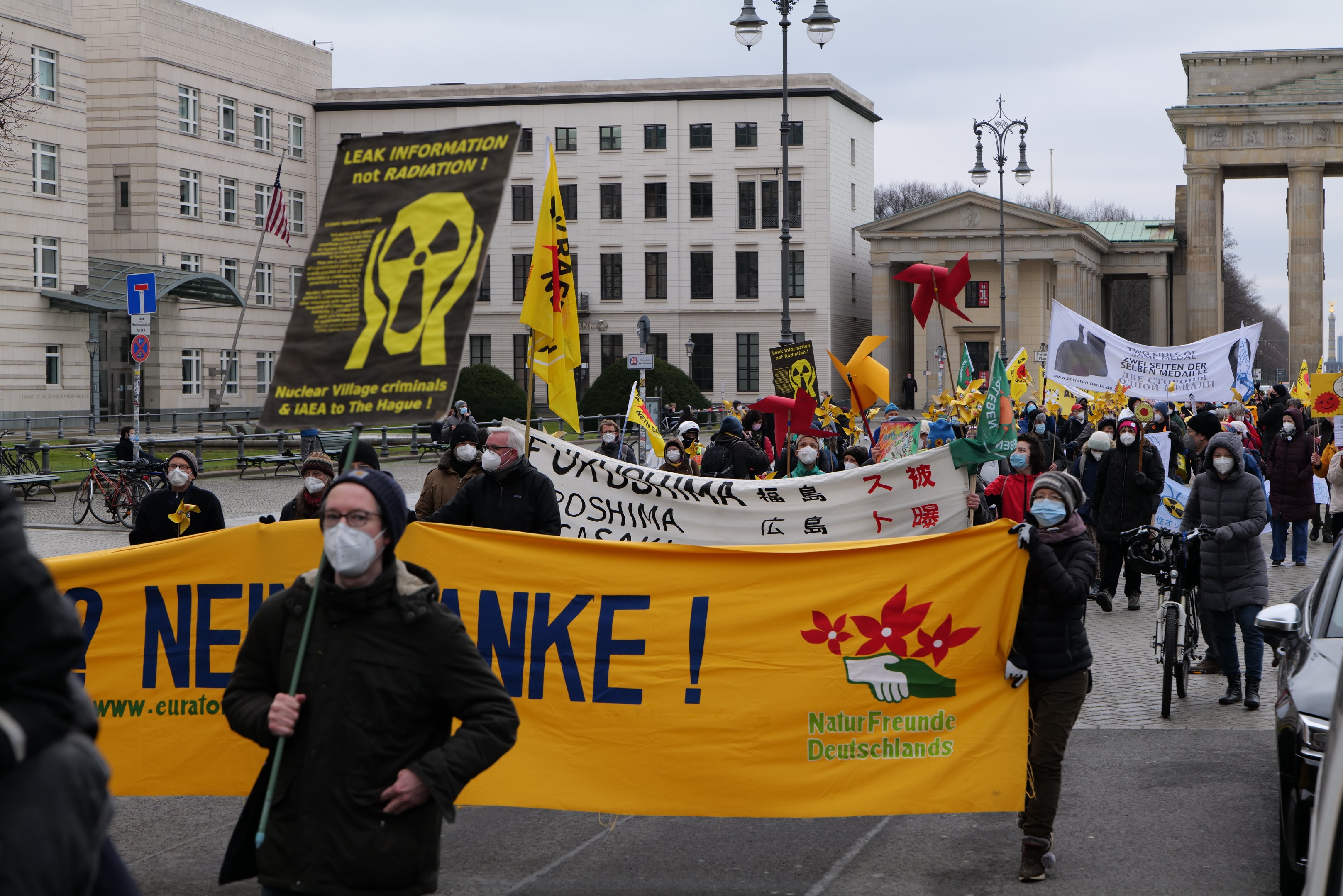Eine große Gruppe von Menschen marschiert mit Schildern und Fahnen auf einer Straße bei einer Demonstration gegen Atomkraft in Deutschland, mit Fahrzeugen auf der rechten Seite und Gebäuden, Laternen, Bäumen und einem Bogen im Hintergrund.
