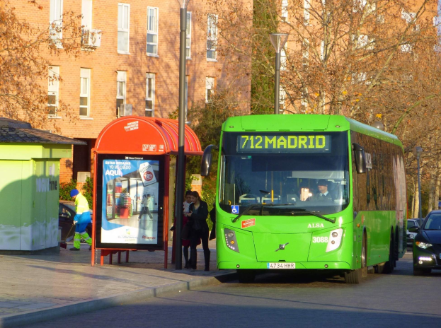 Ein grüner Bus fährt auf einer Straße mit hohen Gebäuden, Fußgängern auf dem Gehweg, einem Schuppen, Laternenpfählen, Bäumen und einem klaren blauen Himmel im Hintergrund.